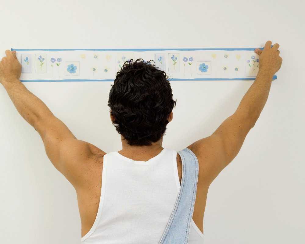 Homem de costas aplicando uma faixa de papel de parede com estampa floral azul em parede branca